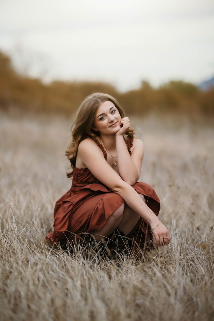 Girl sitting in tall grass wearing a rust dress and cowboy boots